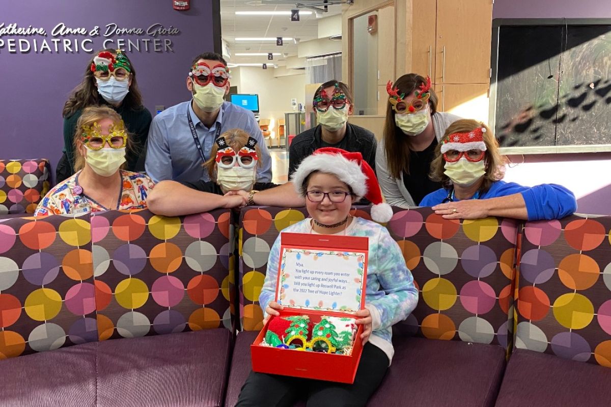 A young girl wears a Santa hat and holds a box of fuzzy slippers. Behind her are nurses wearing masks and funny glasses with Christmas-themed characters. 