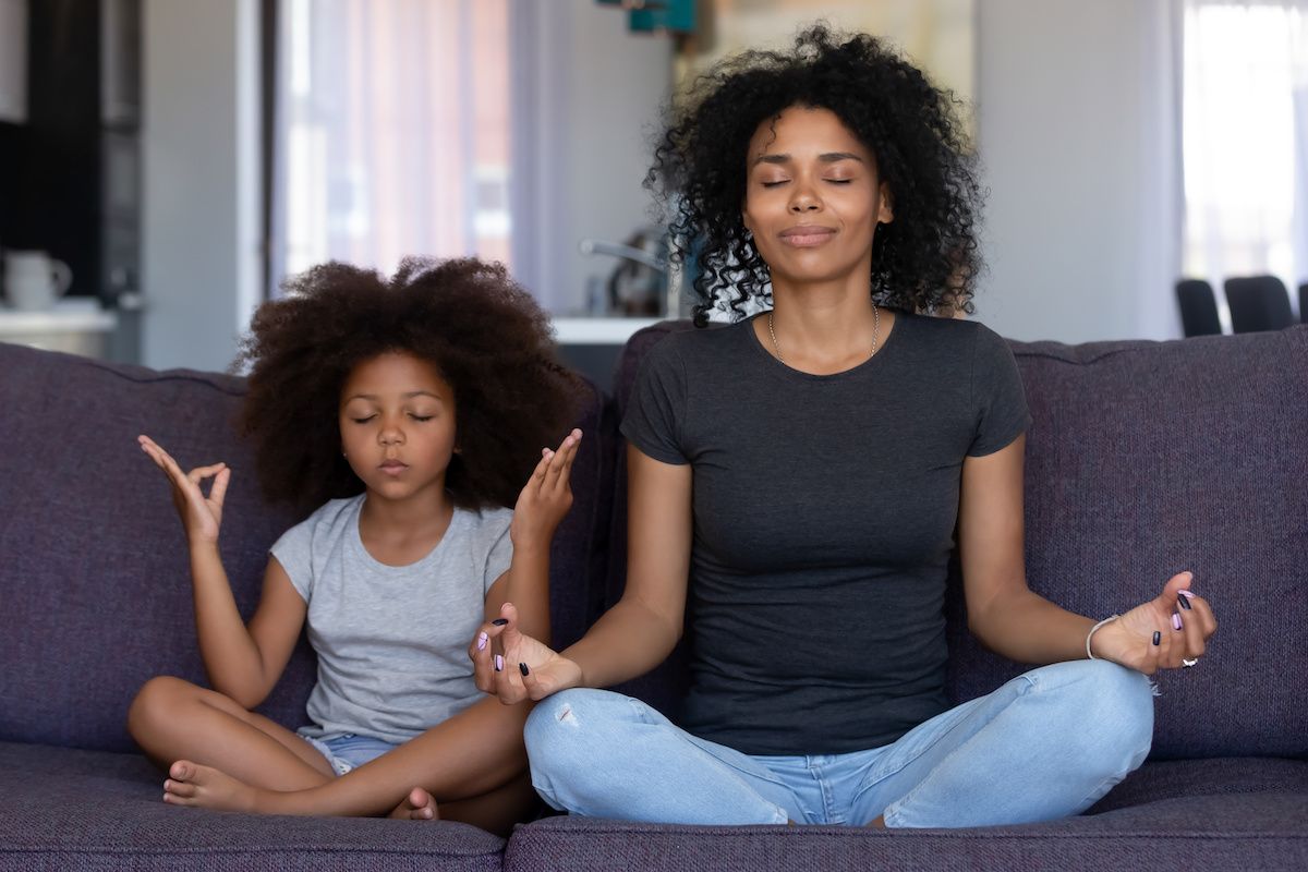 A mother and her young daughter meditating in the yoga lotus position on a sofa