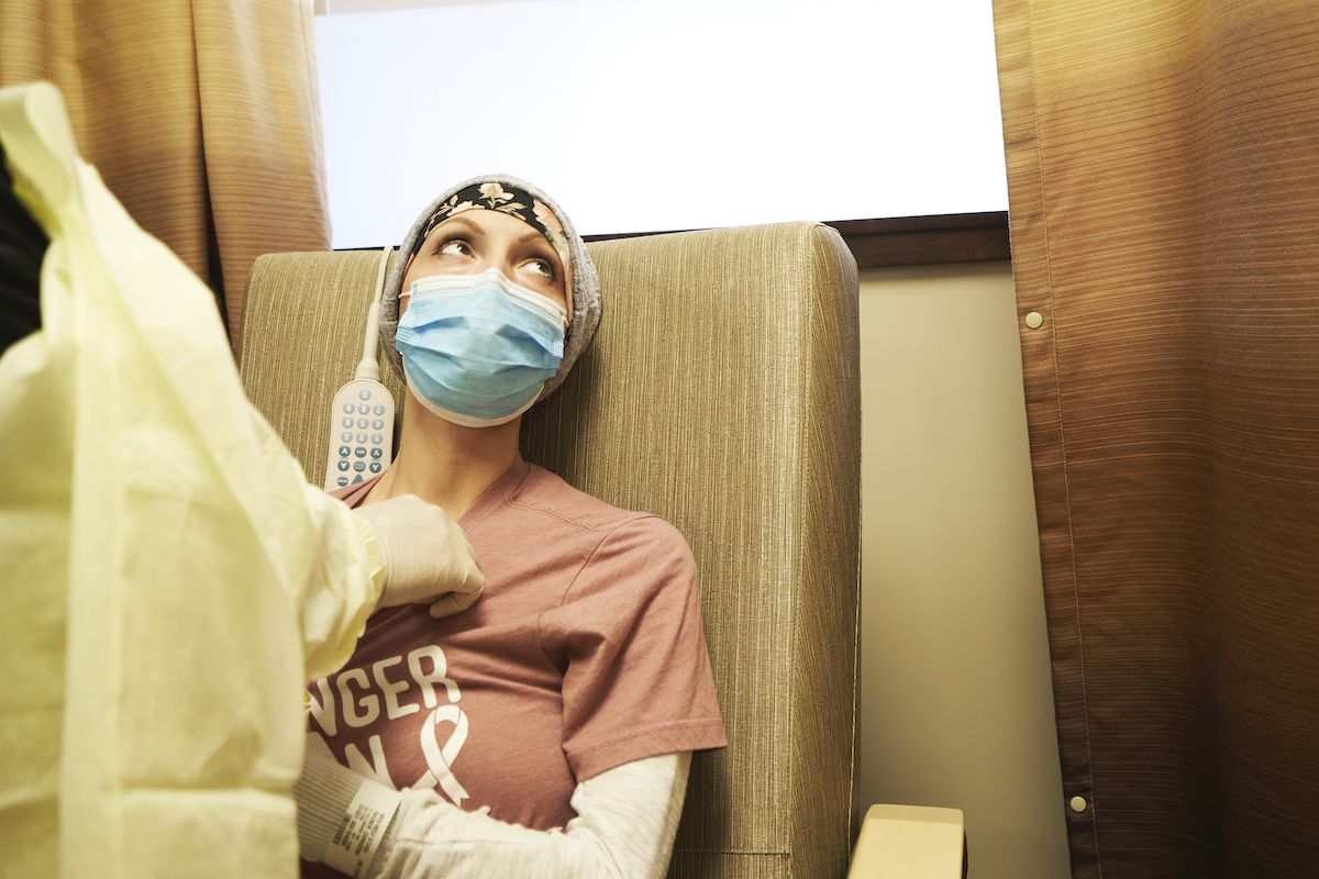 Woman wearing medical mask sits in chair waiting for chemotherapy to begin.
