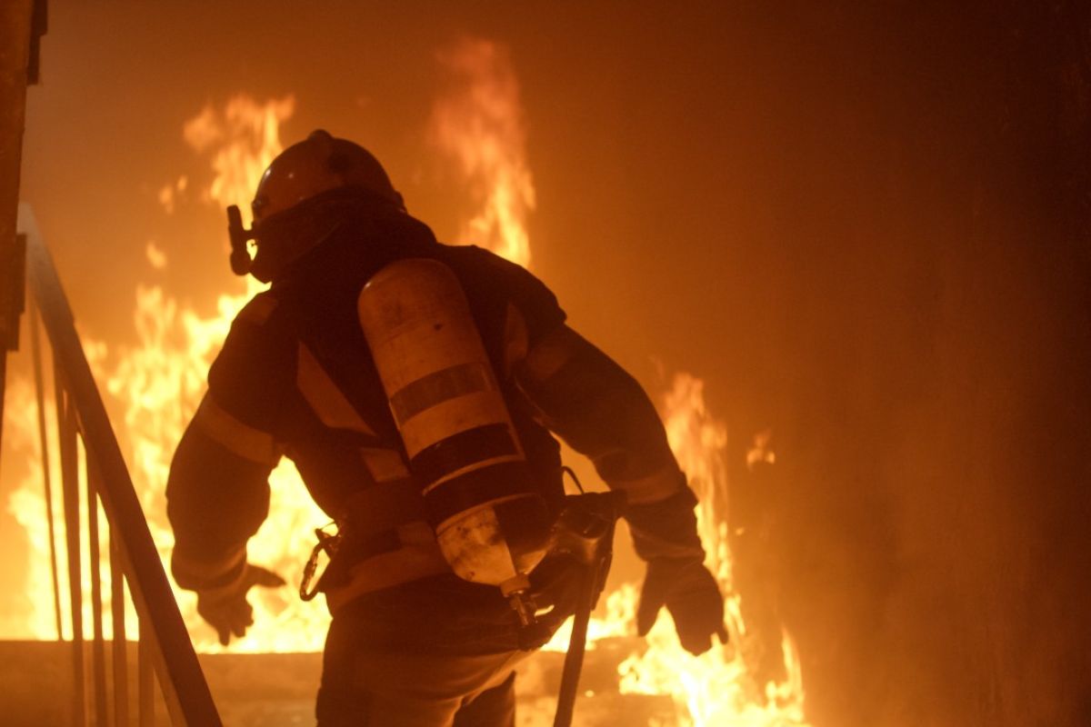 A firefighter in full protective gear walks up stairs into a poorly lit burning building. 