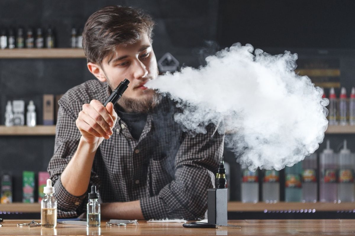 Young adult man holding a vape and blowing smoke while standing behind a bar. 