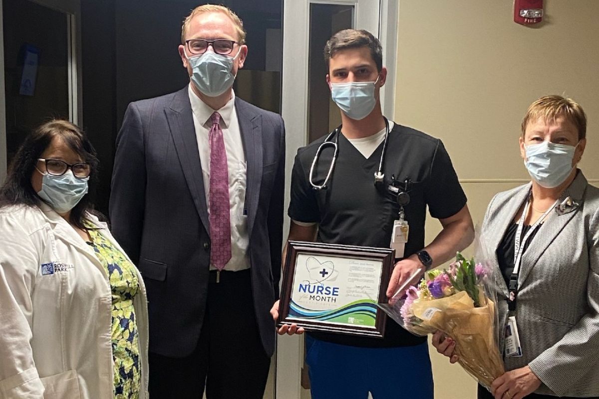 Four masked medical professionals stand together, with the second man, standing second from right, holding a certificate indicating he's been selected as Nurse of the Month for April. 