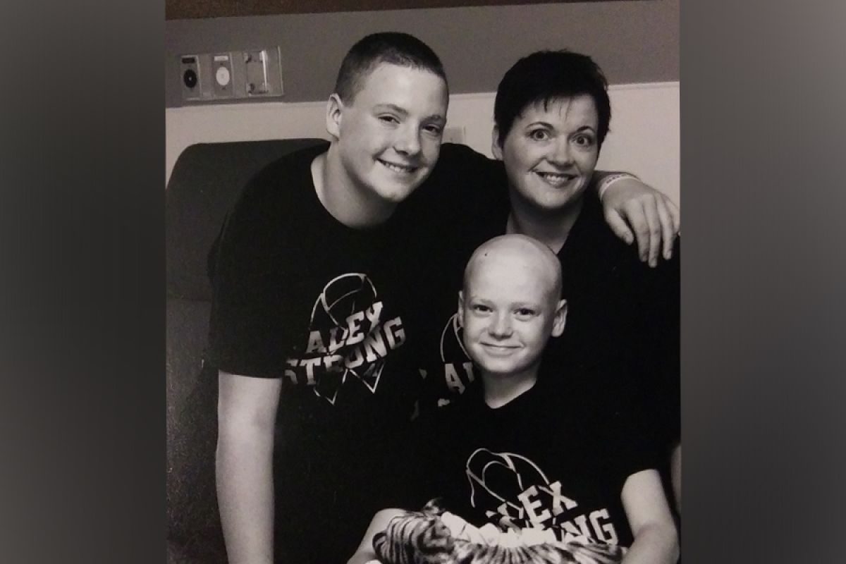 A black and white photo of a mother and her two sons sitting on a hospital bed, wearing shirts that say 'Alex Strong' in honor of the youngest boy in the photo who is fighting cancer.