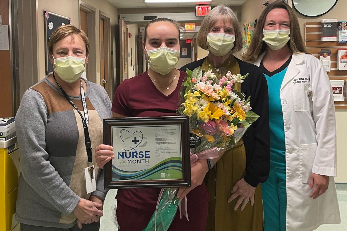 Four women stand in a hospital hallway; the woman second from left holds a bouquet of flowers and a certificate for being named Nurse of the Month for November. 