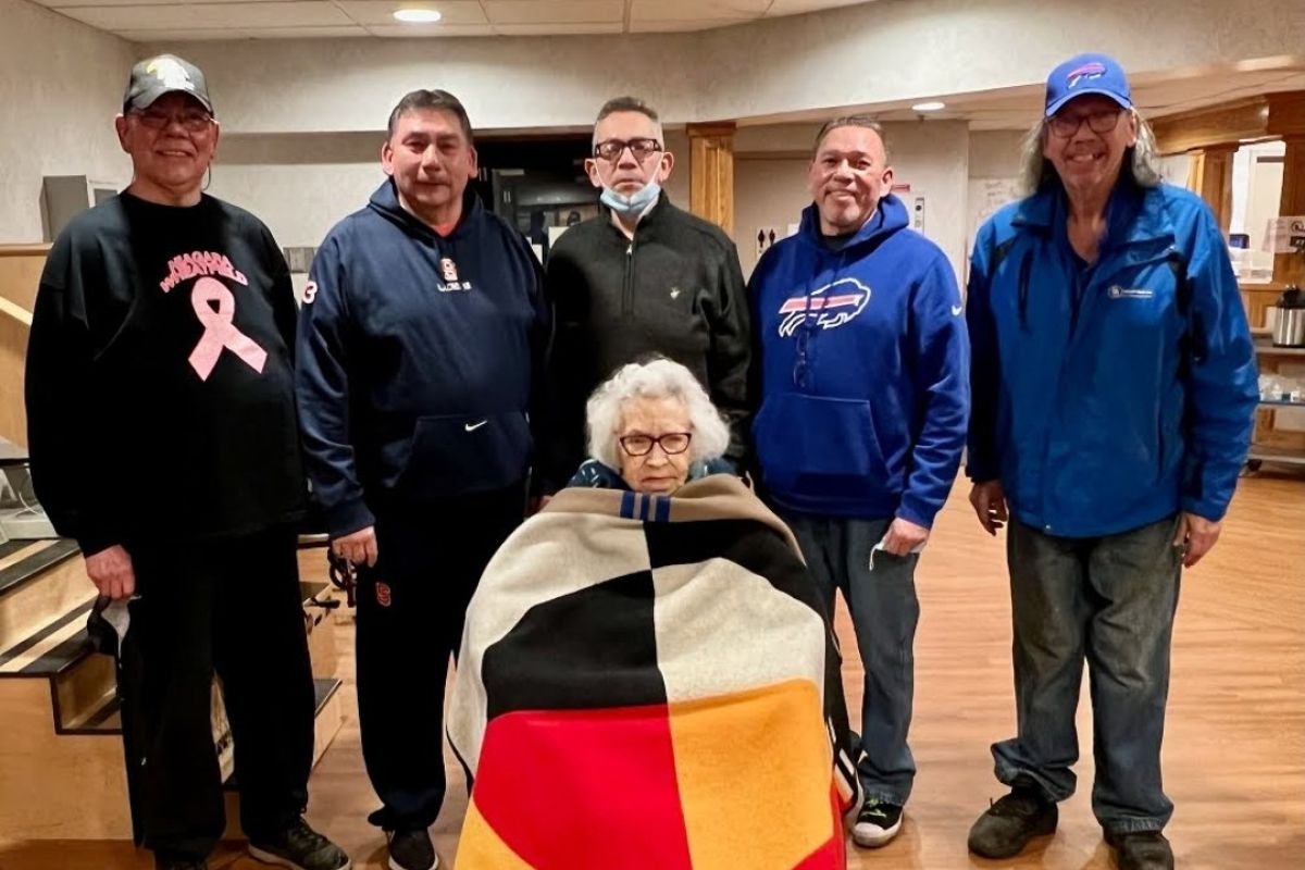 Carolyn Henry sits in a wheelchair, covered by a blanked with red, black, white and yellow segments, in front of her sons (L-R) Dale, Rob (Bear), Ron (Mouse), Brian and Ray. 