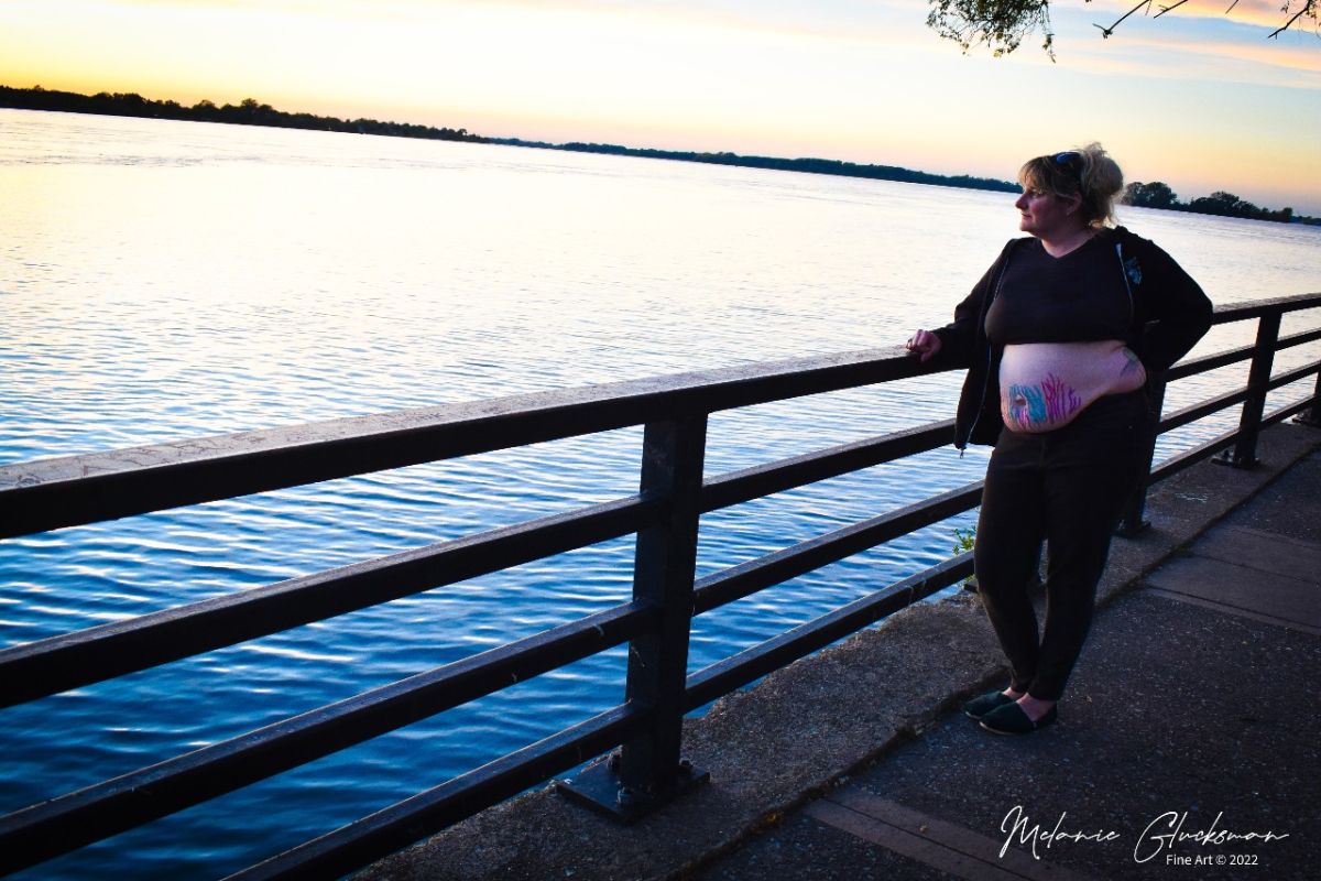 A woman stands on a sidewalk overlooking a river, with her stomach exposed and a row of multicolored glitter stripes on her abdomen. 