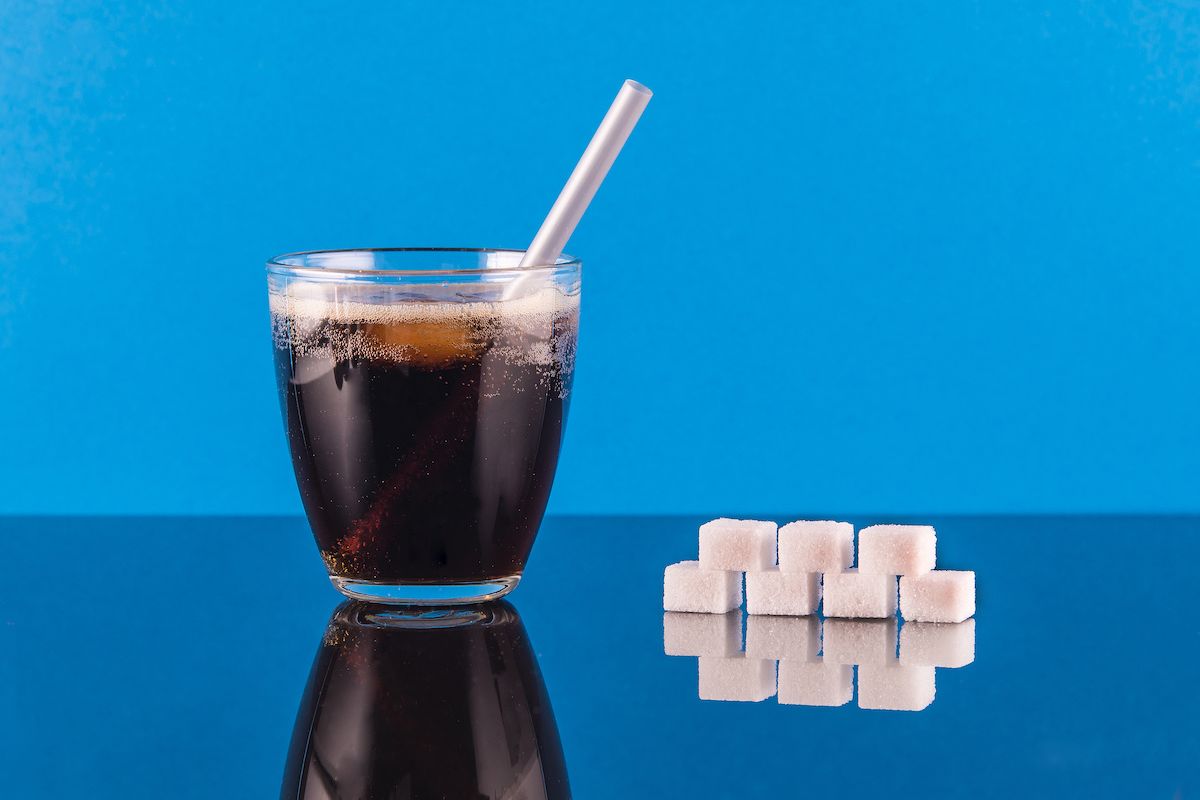 A glass of pop and a pile of sugar cubes sits on a table with a blue background