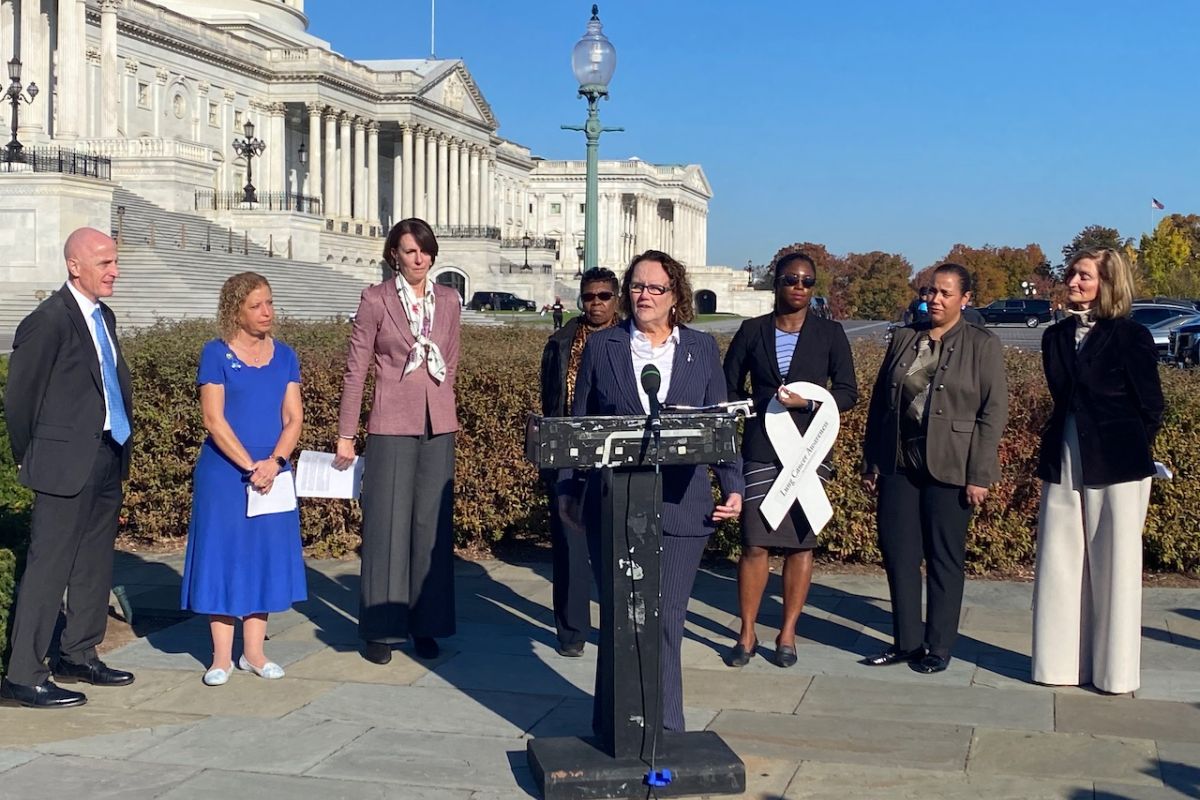 A woman in a suit speaks at a podium near Capitol Hill with others standing around her