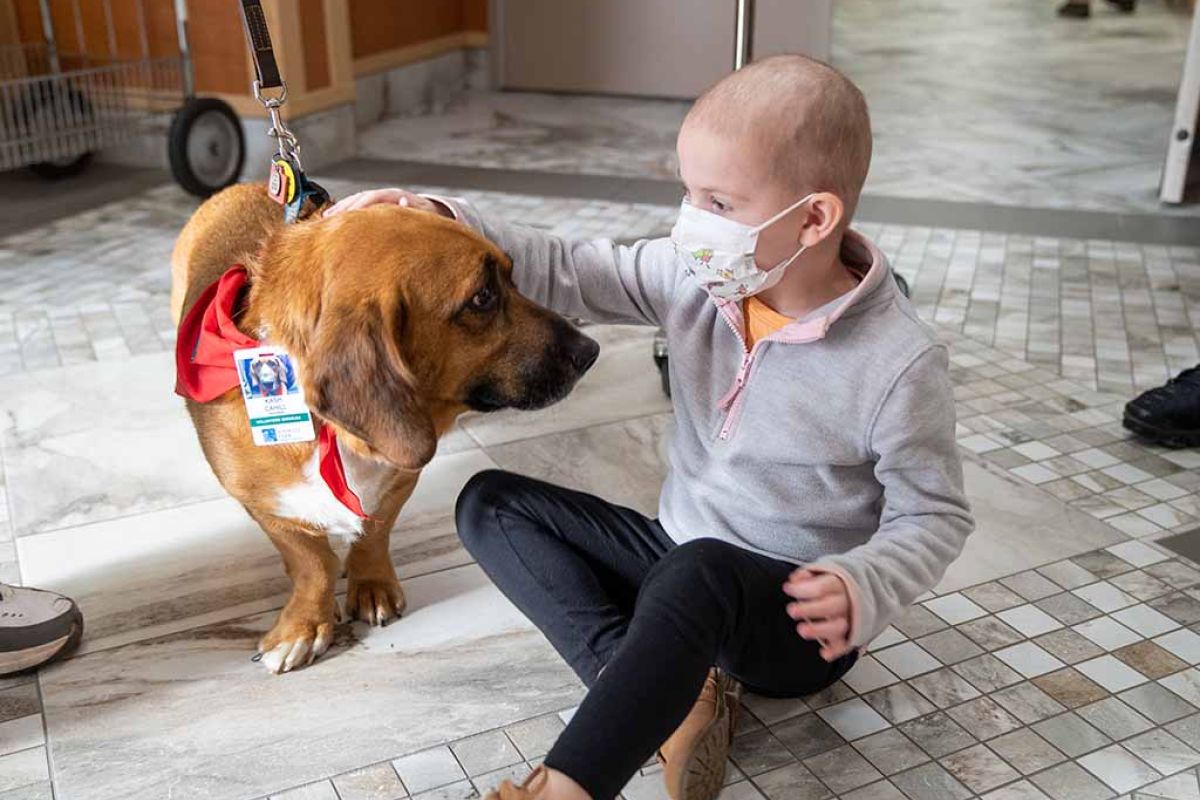 A young girl sits on the floor of a clinic petting a therapy dog. 