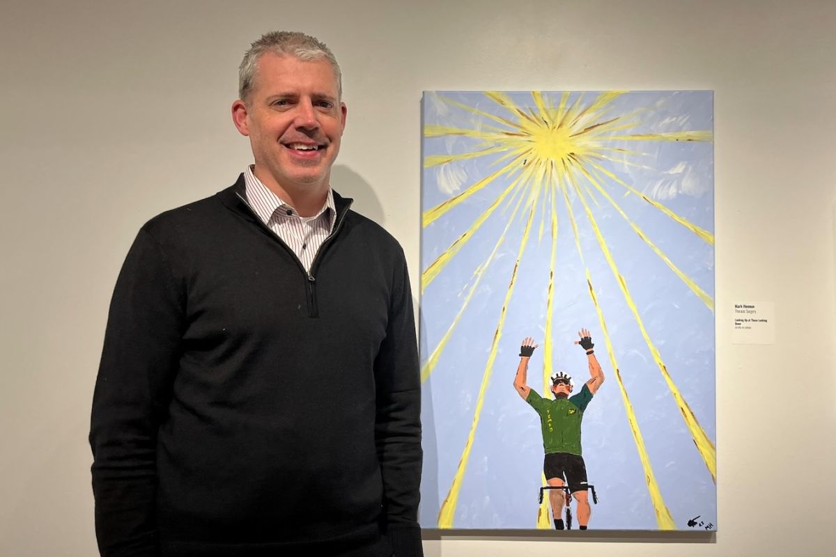 Dr. Mark Hennon poses with his painting in the Art Heals Gallery for the 2025 Employee Art Show, featuring a cyclist looking up at the sky.