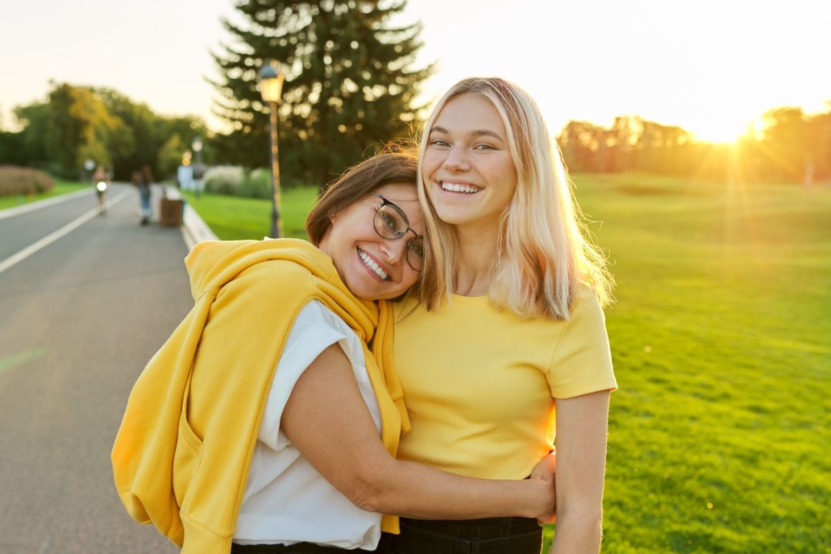 A women hugs her teenager daughter and smiles at the camera in the park