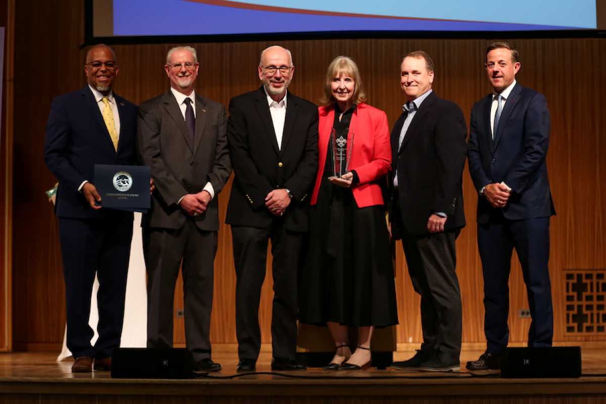 Woman in red jacket receives award next to five men