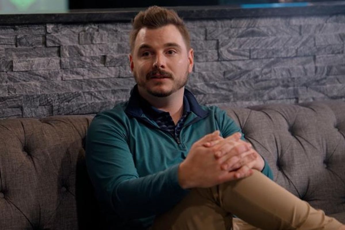 Man sits on a couch with leg crossed, talking to someone sitting to his side. 