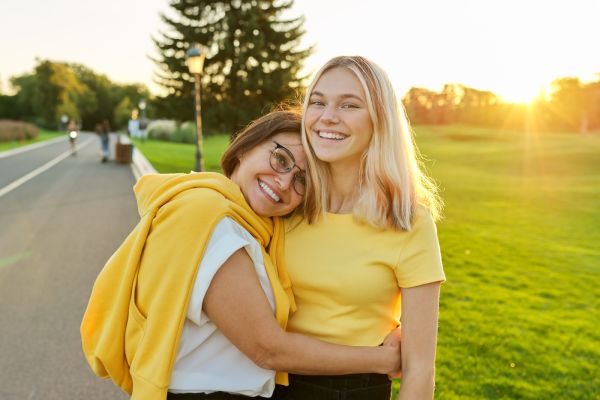 A women hugs her teenager daughter and smiles at the camera in the park