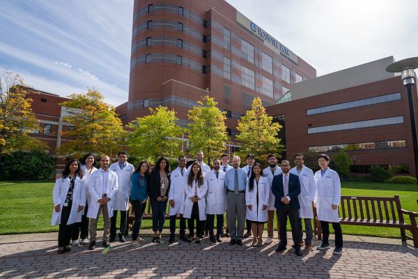 A group of current fellows, graduates and program leads of the Hematology Oncology fellowship program smile in a group in Kaminski Park with Roswell Park in the backgrounc