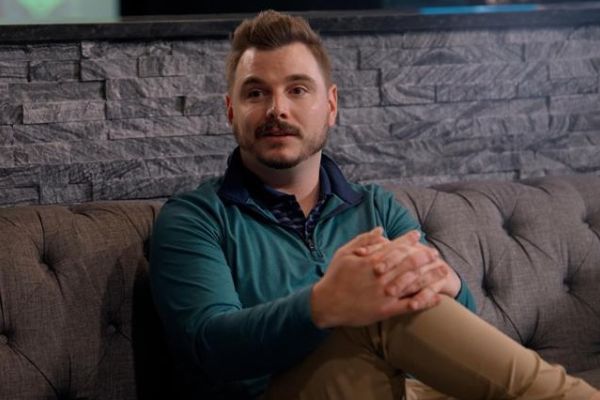 Man sits on a couch with leg crossed, talking to someone sitting to his side. 