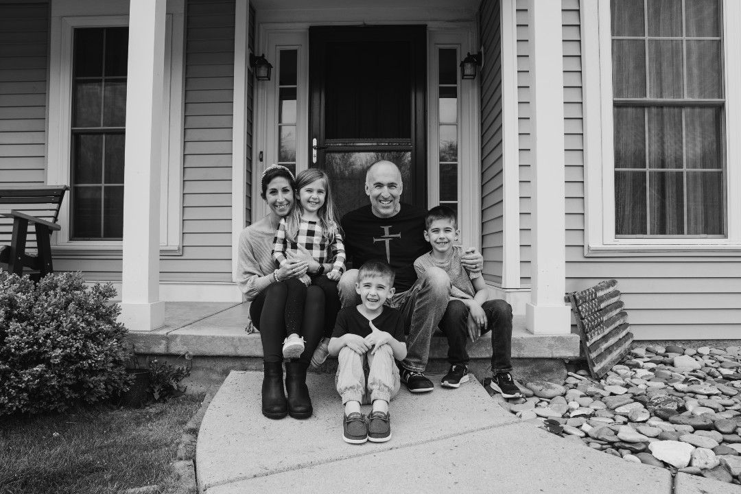 A black and white photo of a family of five on the front porch of a house, mother is holding daughter on her lap, one son is sitting on the sidewalk and the father has his arm around the other son's shoulder. 