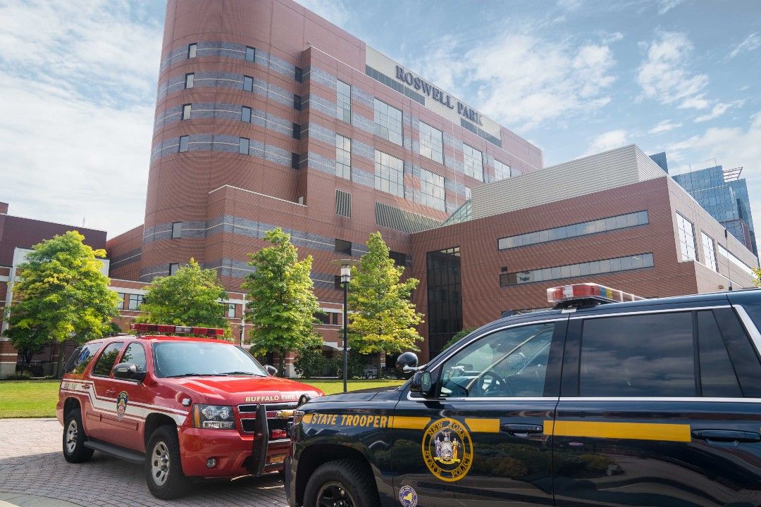 A red Buffalo Fire Department SUV is parked in front of a blue New York State Trooper SUV with Roswell Park's main hospital building in the background.