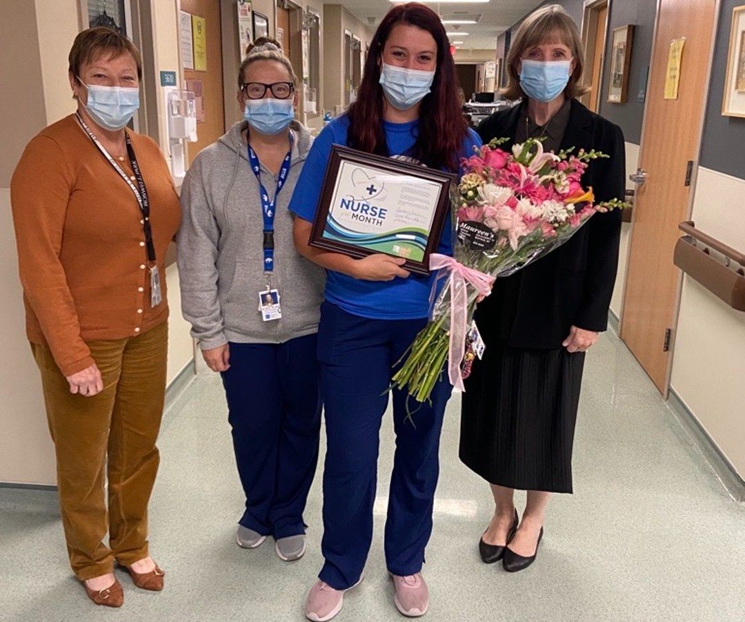 A group of four women, wearing surgical masks, stand in a hallway. The woman in the middle is holding flowers and a certificate for being named Nurse of the Month for October. 