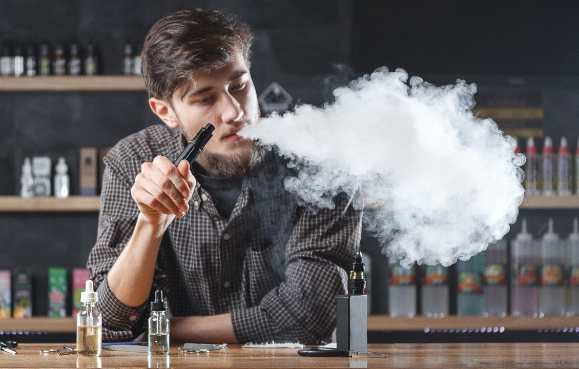 Young adult man holding a vape and blowing smoke while standing behind a bar. 