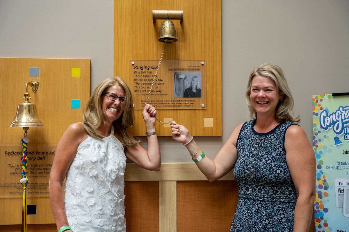 Two women stand together, each holding a piece of the string used to ring the Victory Bell after completing cancer treatment. Both women are smiling proudly and laughing. 