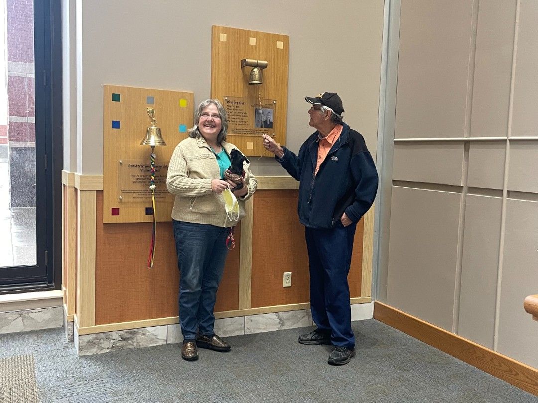 A man stands in front of the Victory Bell, wearing a dark coat and baseball cap, while his wife and their dog cheer him on. 