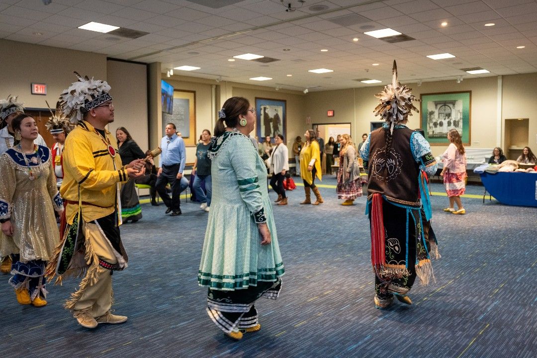 A group of dancers, wearing traditional Indigenous costumes, and staff members from Roswell Park participate in traditional dances as part of a Native American Heritage Month celebration.