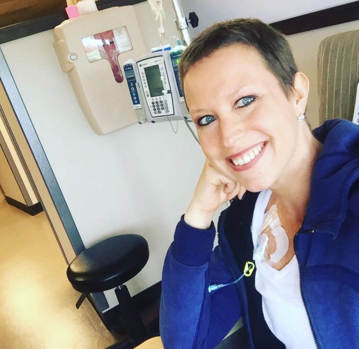 A woman with very short dark hair, wearing a blue coat, smiles at the camera from a chair while receiving chemotherapy. 