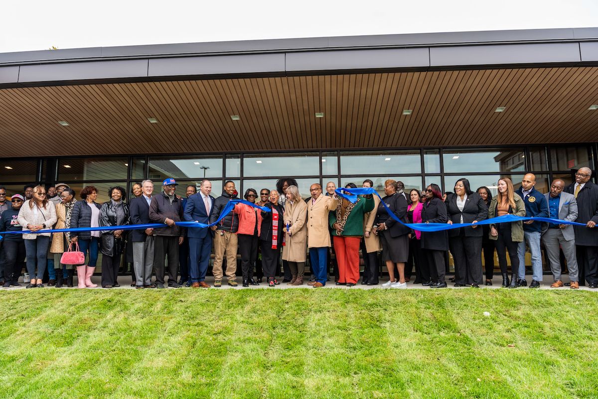 People gathered to cut a blue ribbon in front of a building, with grass in foreground