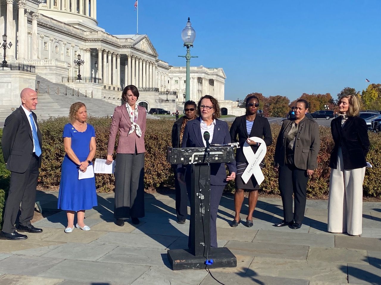 A woman in a suit speaks at a podium near Capitol Hill with others standing around her