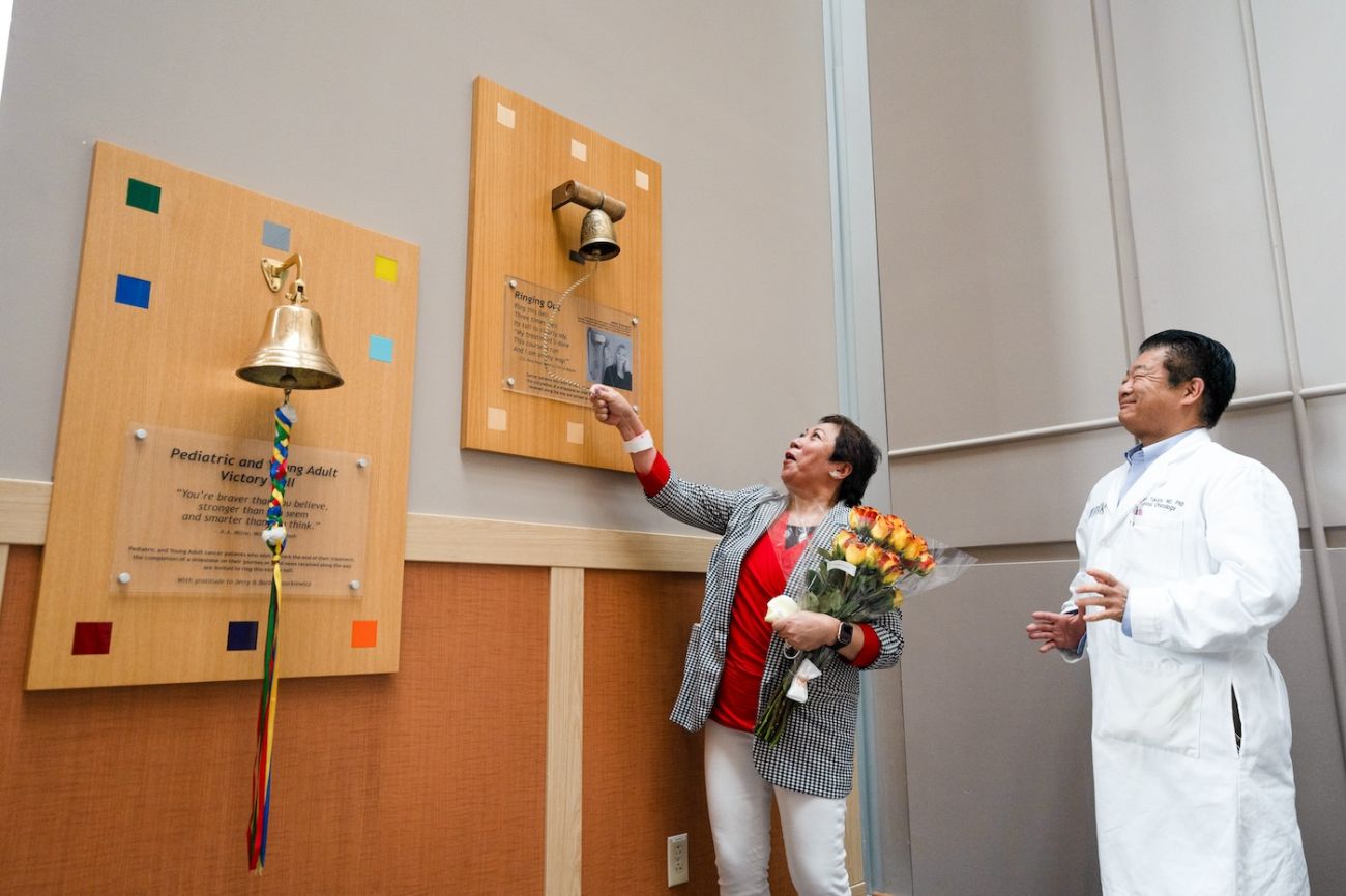 Edith Lazatin rings the victory bell, holding a bouquet of flowers, while Dr. Kazuaki Takabe cheers her on.
