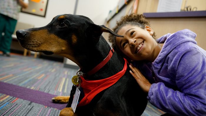 A young girl sits on the floor of a clinic hugging a therapy dog. 
