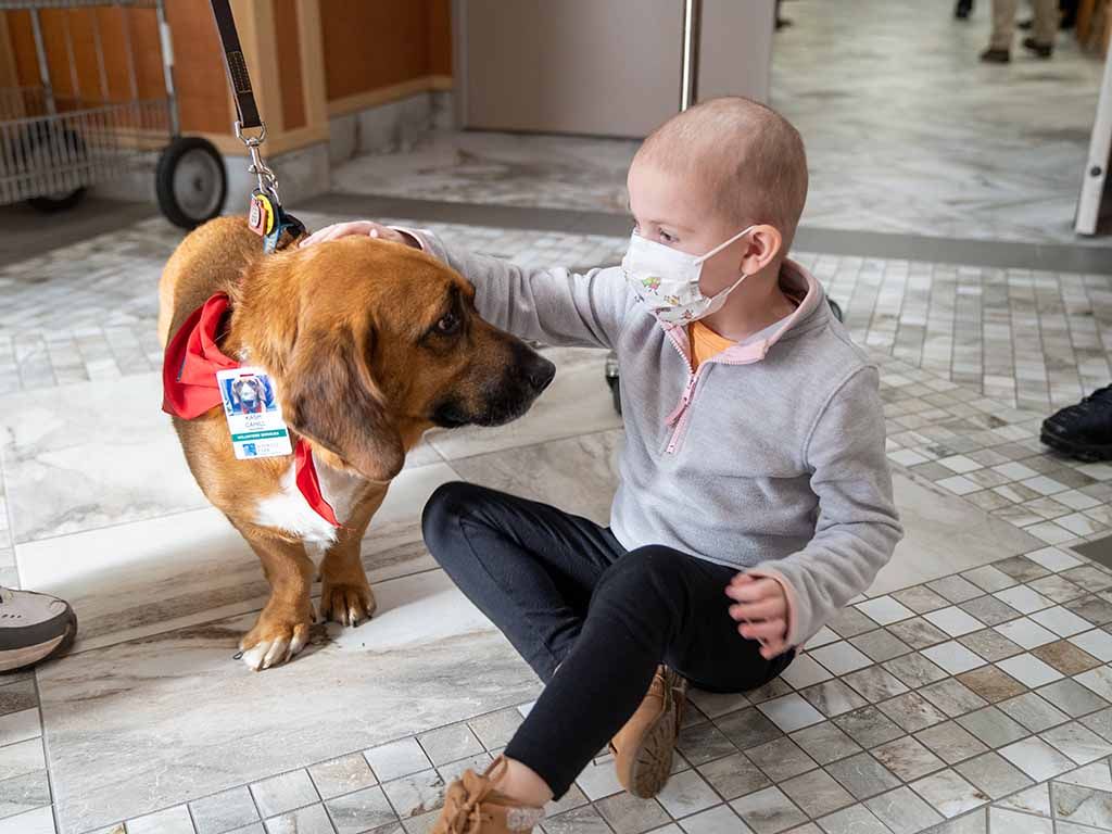 A young girl sits on the floor of a clinic petting a therapy dog. 