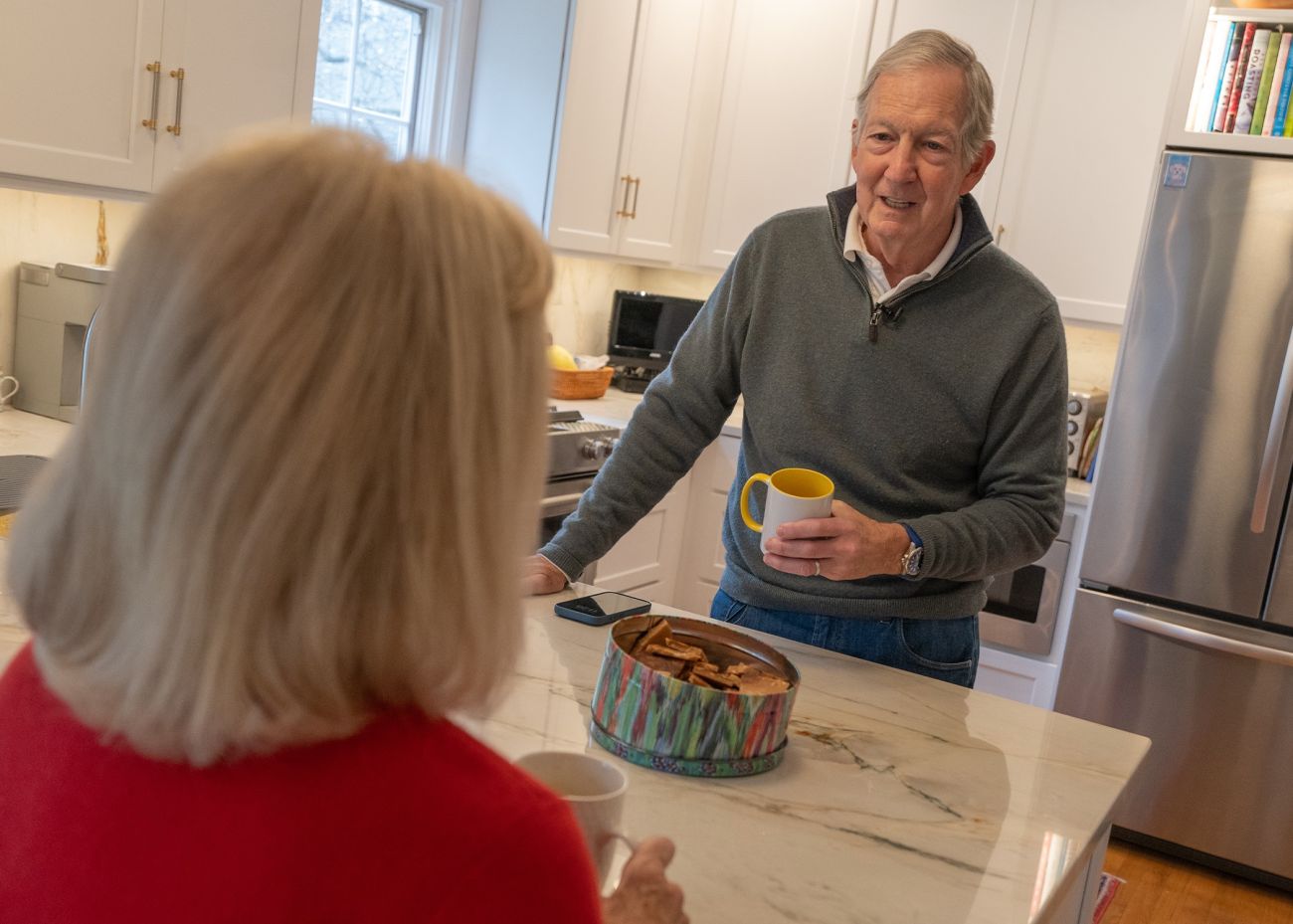 A grey-haired man in a blue sweater holds a coffee cup as he talks with a woman in a kitchen