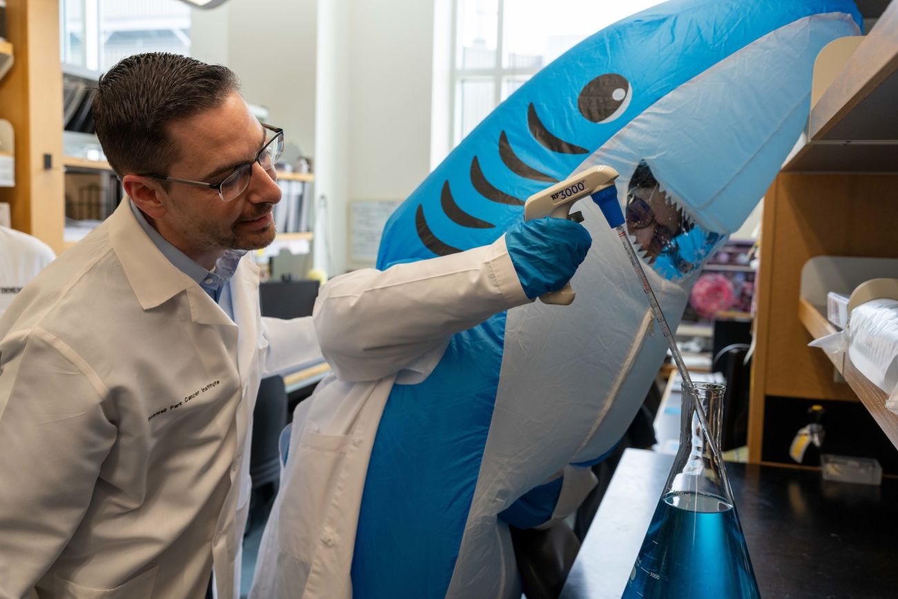 A scientist works at a lab bench with a person in a shark costume
