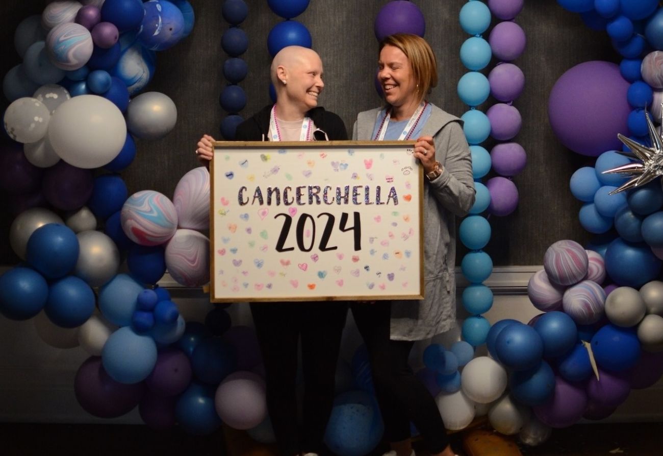Two women hold a sign that says "Cancerchella 2024" in a photobooth at the Young Adult Retreat 