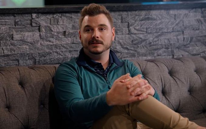 Man sits on a couch with leg crossed, talking to someone sitting to his side. 