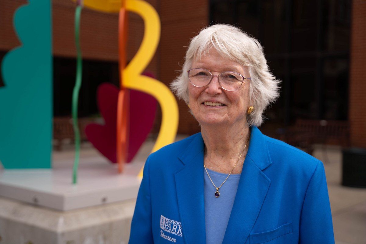 A white-haired woman wears a blue Roswell Park volunteer and stands in front of a sculpture. 
