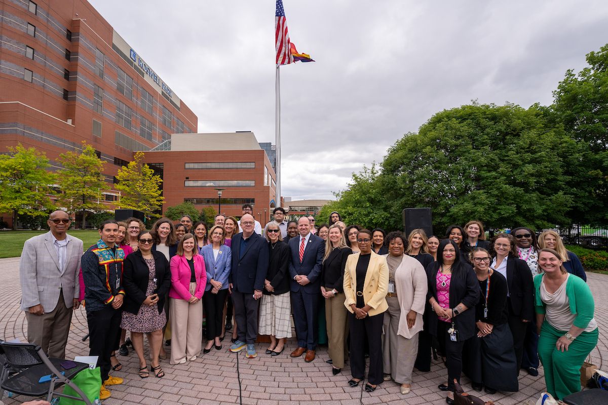 A group of people stands gathered around a flagpole with a hospital behind them