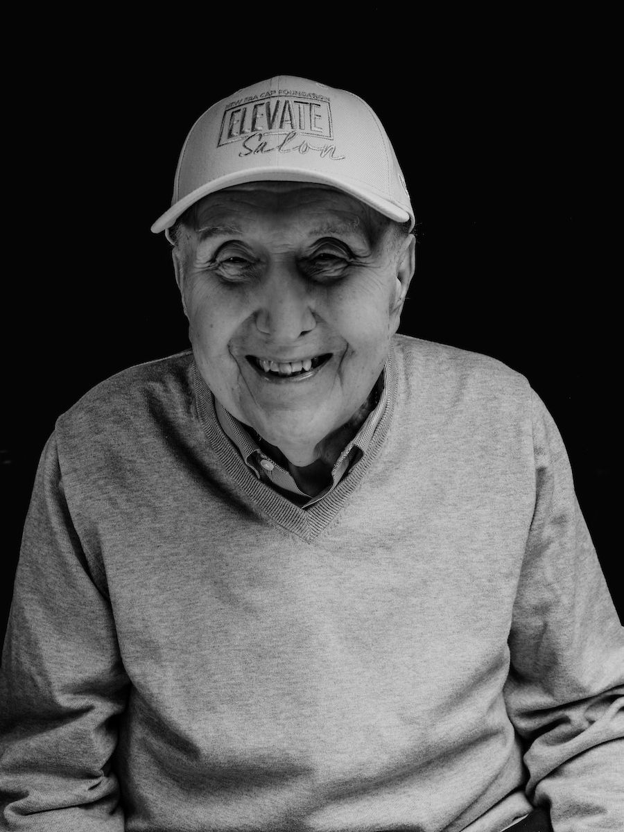 Black and white photo of metastatic breast cancer patient Joe Gadawski poses with a hat that reads Elevate Salon