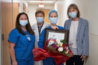 Four medical professional women standing in a hallway with masks; one is holding flowers and a certificate for being Roswell Park's Nurse of the Month for June. 