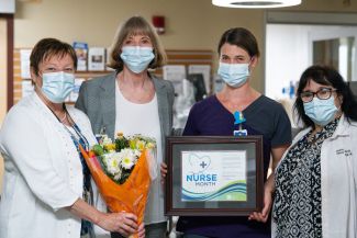 Four medical professional women standing in a hallway with masks; one is holding flowers and a certificate for being Roswell Park's Nurse of the Month for July. 