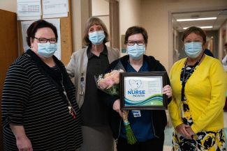Four medical professional women standing in a hallway with masks; one is holding flowers and a certificate for being Roswell Park's Nurse of the Month for August. 