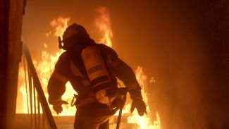 A firefighter in full protective gear walks up stairs into a poorly lit burning building. 