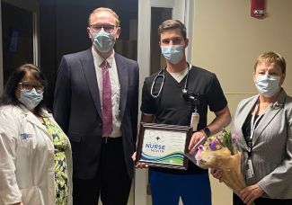 Four masked medical professionals stand together, with the second man, standing second from right, holding a certificate indicating he's been selected as Nurse of the Month for April. 