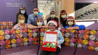 A young girl wears a Santa hat and holds a box of fuzzy slippers. Behind her are nurses wearing masks and funny glasses with Christmas-themed characters. 