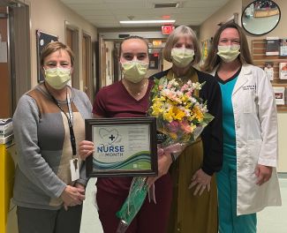 Four women stand in a hospital hallway; the woman second from left holds a bouquet of flowers and a certificate for being named Nurse of the Month for November. 