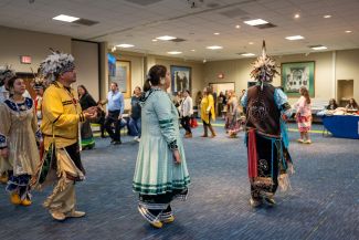 A group of dancers, wearing traditional Indigenous costumes, and staff members from Roswell Park participate in traditional dances as part of a Native American Heritage Month celebration.