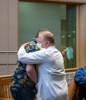 Two men hug, one is wearing a lab coat and a surgical mask. 