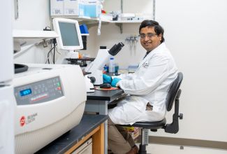 A man with dark hair in a white lab coat smiles in a research lab